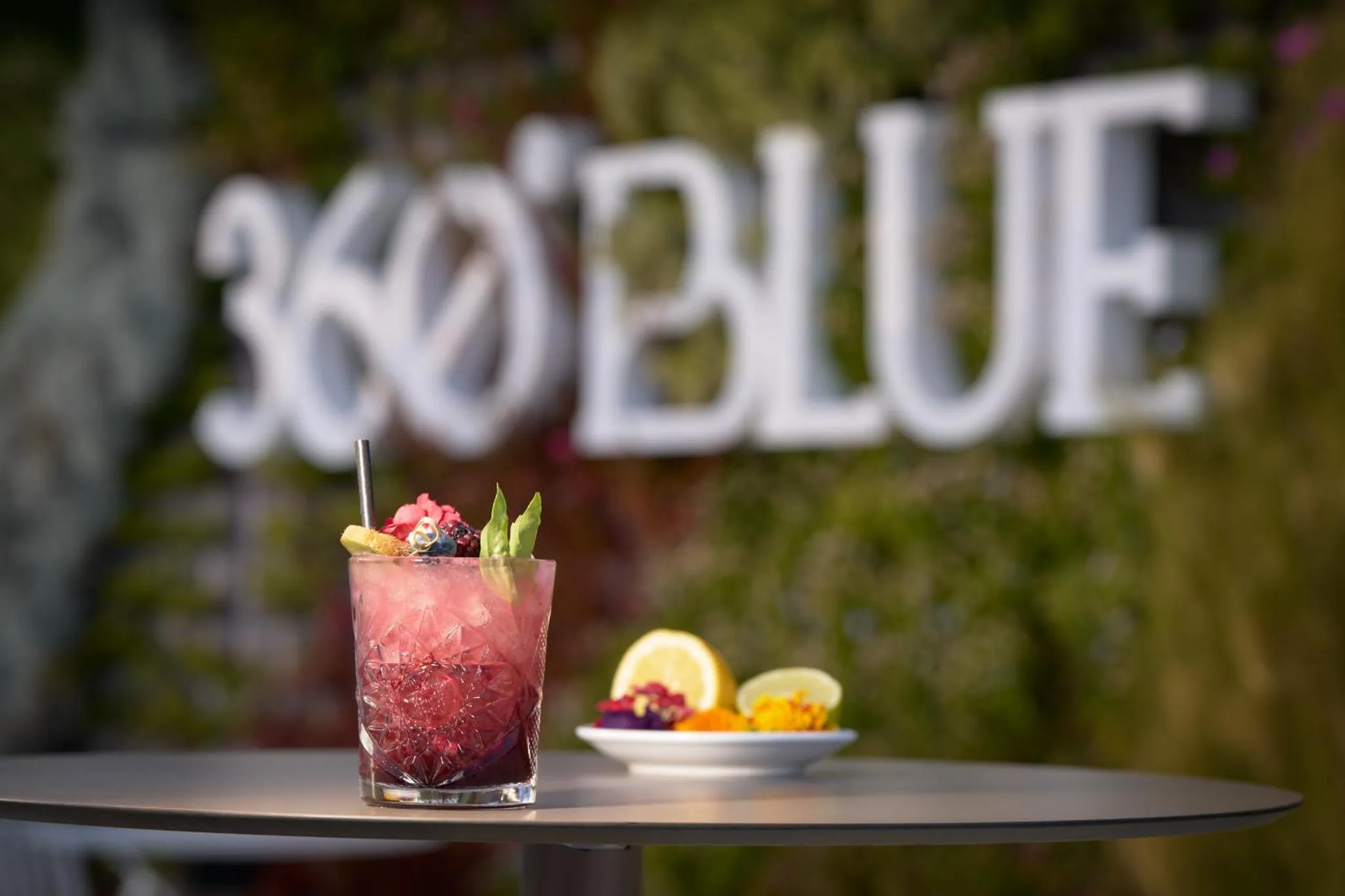 Un cocktail coloré et des amuse-gueules sont présentés sur une table sombre devant un décor lumineux.