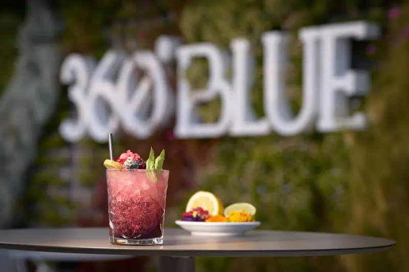 Un cocktail coloré et des amuse-gueules sont présentés sur une table sombre devant un décor lumineux.