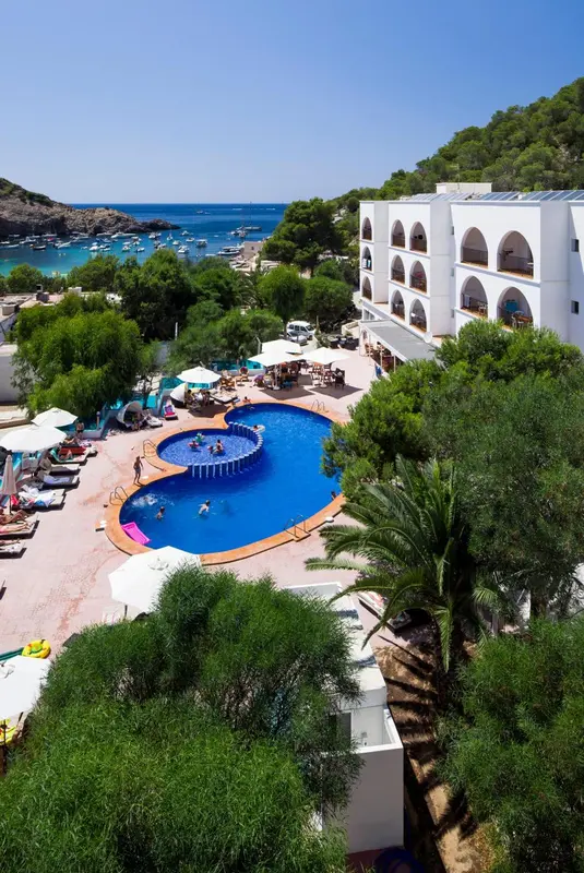 Vue aérienne d'un hôtel blanc avec piscine et végétation luxuriante surplombant une crique.