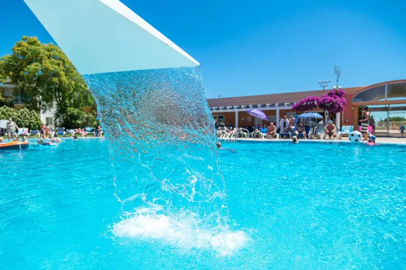 Une cascade d'eau se déverse dans une piscine turquoise entourée de chaises longues et de parasols.