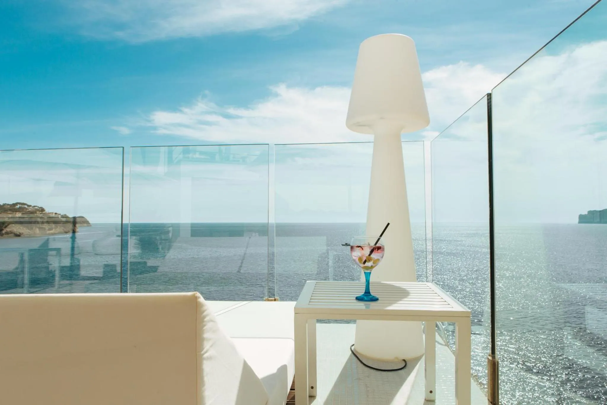 Une table basse avec un verre coloré se trouve sur une terrasse lumineuse avec vue sur la mer.