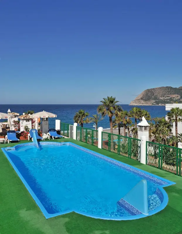 Une piscine rectangulaire turquoise avec un toboggan, entourée d'une pelouse artificielle et de palmiers, surplombe la mer.