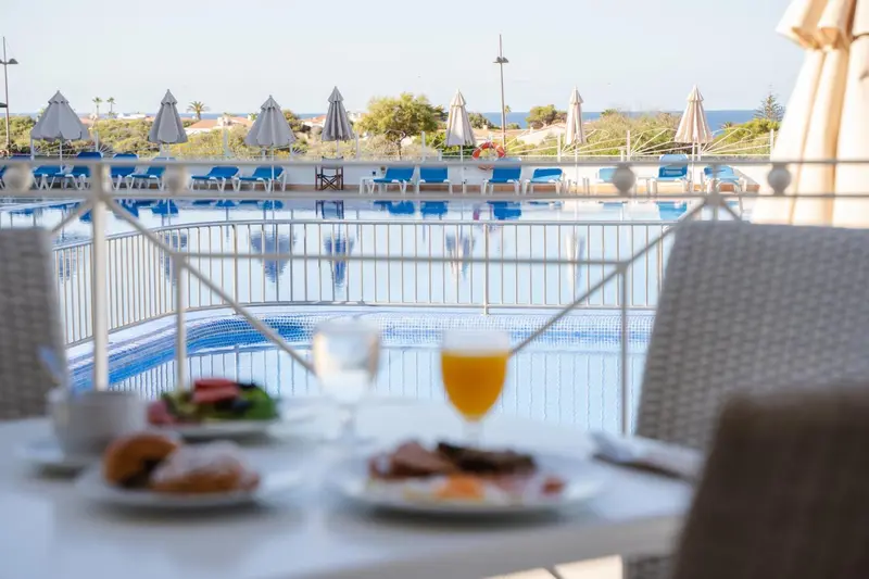 Une table est dressée avec un petit-déjeuner devant une piscine ensoleillée.