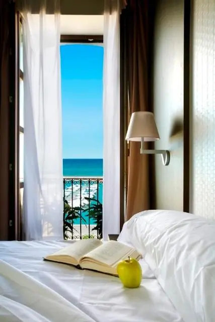 Un lit avec un livre ouvert et une pomme verte, devant une fenêtre ouverte sur la mer.