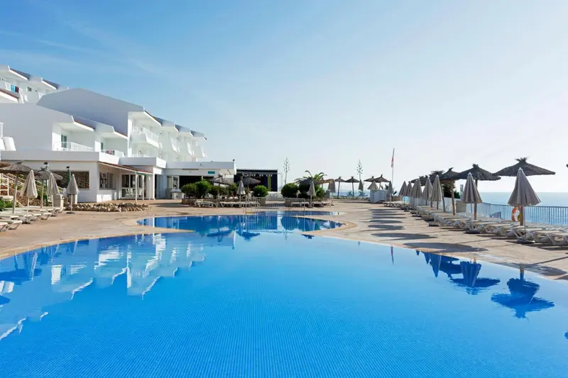 Une piscine bleue reflète un bâtiment blanc et des parasols sur une terrasse ensoleillée.