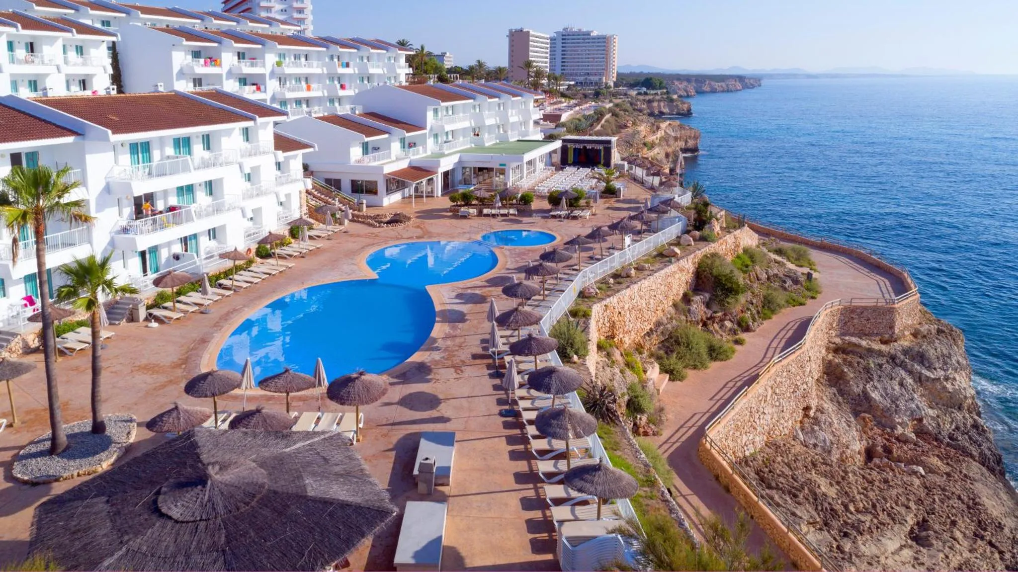 Vue aérienne d'un complexe hôtelier blanc avec des piscines et des transats au bord d'une falaise surplombant la mer.