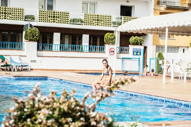 Une femme souriante est assise au bord d'une piscine ensoleillée, entourée de végétation et de chaises longues.