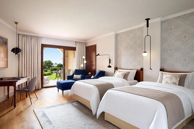 Une chambre lumineuse avec deux lits simples et un fauteuil bleu près d'une porte-fenêtre.