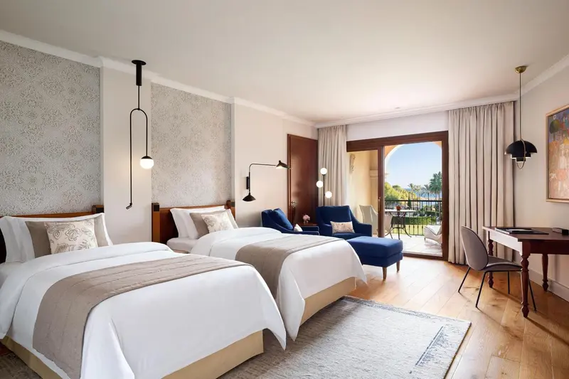 Une chambre lumineuse avec deux lits et un fauteuil près d'une porte-fenêtre.