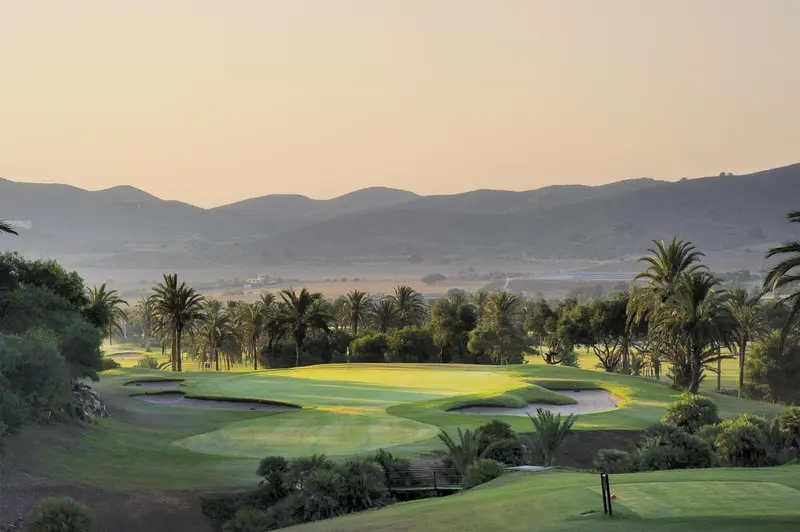 Un terrain de golf verdoyant s'étend devant des collines arides et un ciel orangé.