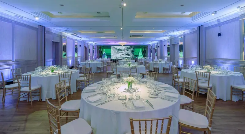 Une salle lumineuse est mise en scène pour un banquet avec des tables rondes nappées et des chaises dorées.