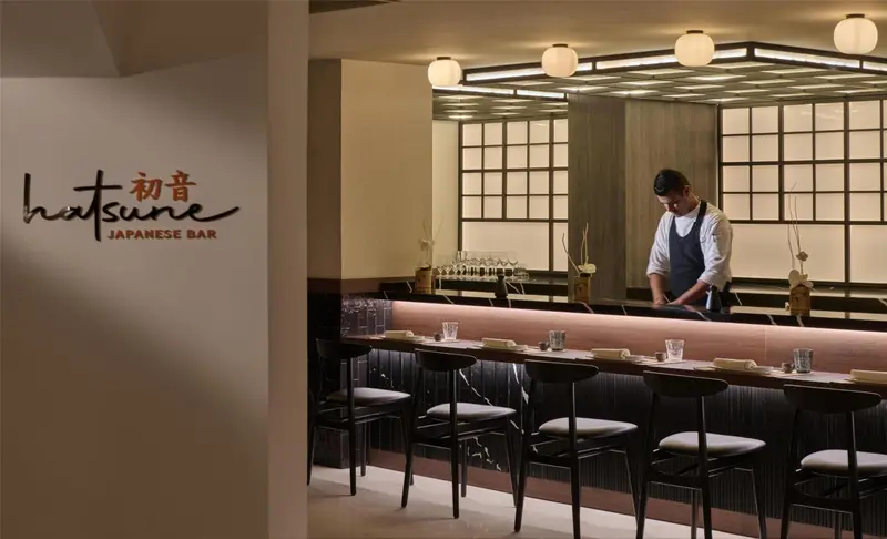 Un comptoir de bar en bois sombre avec des tabourets et un chef préparant des plats.