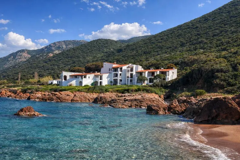 Bâtiment blanc avec toits en tuiles rouges surplombant une plage rocheuse et une mer turquoise.