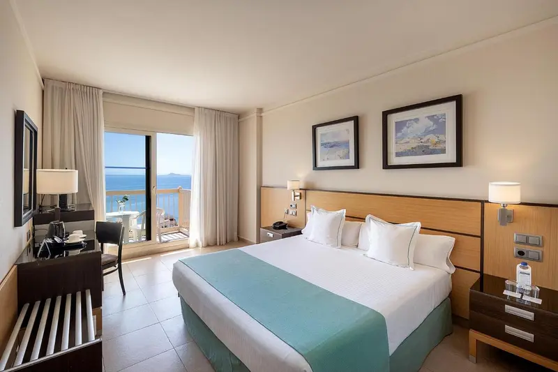 Chambre lumineuse avec un grand lit et une vue sur la mer depuis un balcon.