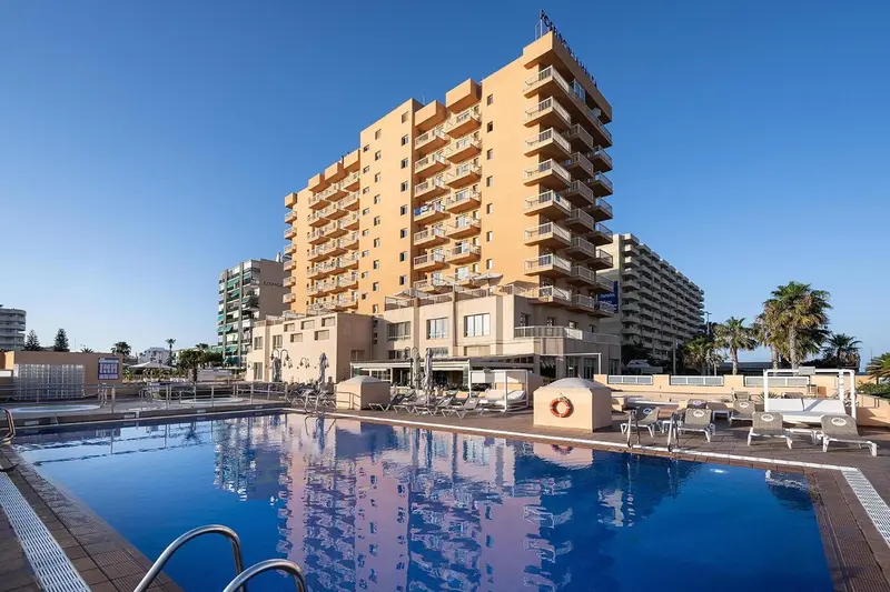 Bâtiment beige à plusieurs étages avec des balcons surplombant une piscine bleu clair.