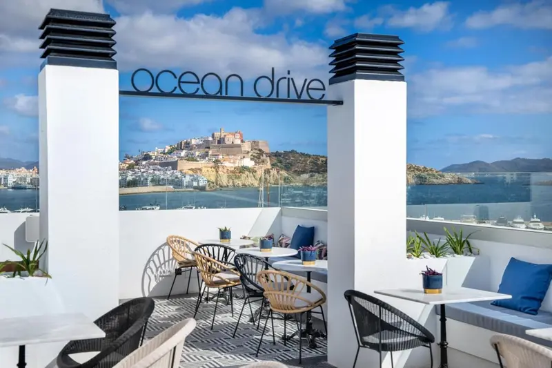 Terrasse aménagée avec tables et chaises offrant une vue sur la mer et une ville côtière.