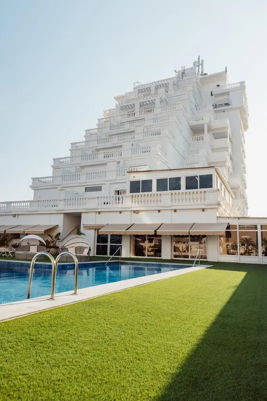 Bâtiment blanc à plusieurs étages avec une piscine au premier plan et une pelouse verte.