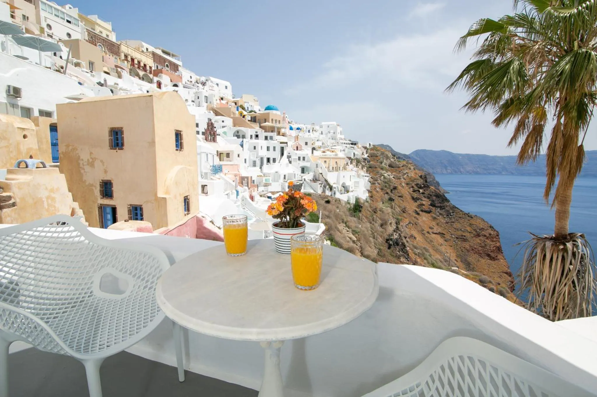 Une table avec des boissons est placée sur une terrasse offrant une vue sur la mer et les bâtiments blancs.