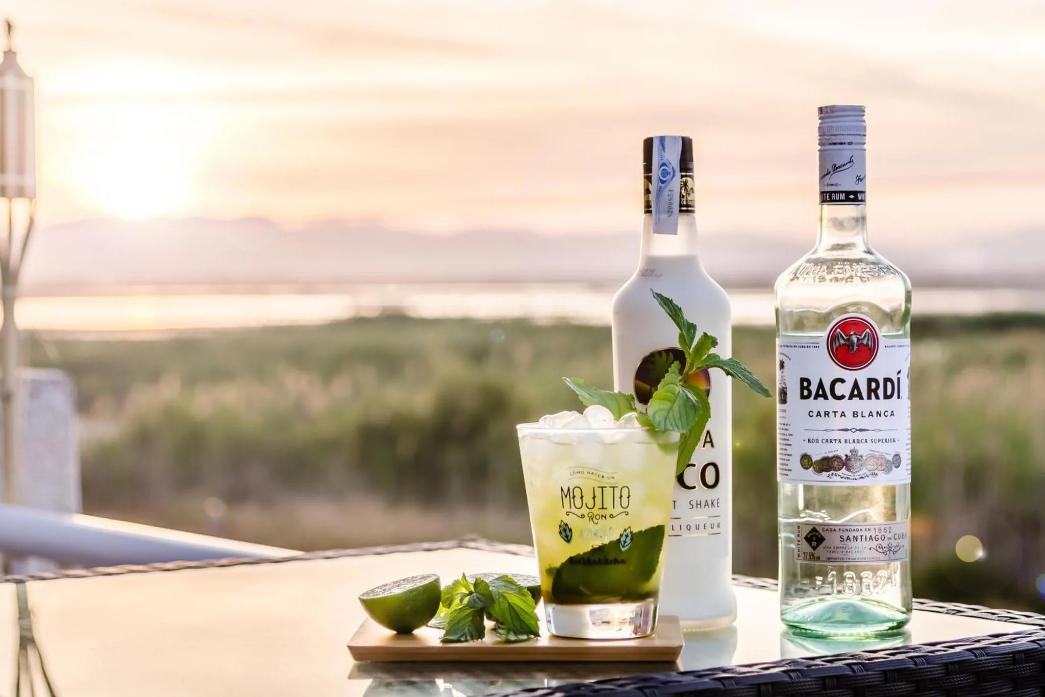 Deux bouteilles de rhum et un cocktail à la menthe sont présentés sur une table en bois au bord d'une terrasse.