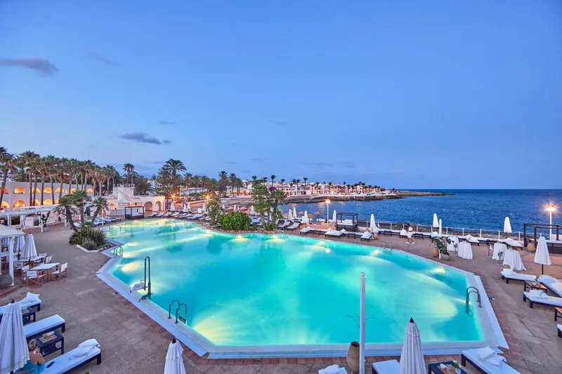 Piscine illuminée d'un établissement hôtelier au bord de la mer, au crépuscule.