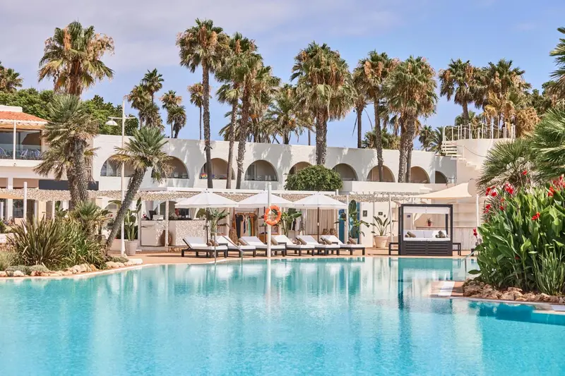 Une piscine turquoise reflète des palmiers et un bâtiment blanc.
