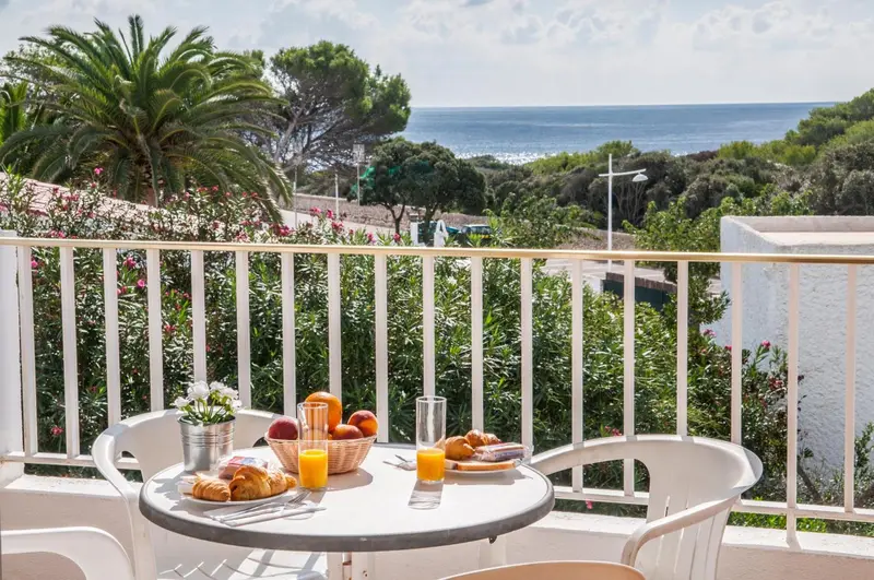Une table ronde avec un petit déjeuner est posée sur une terrasse fleurie avec vue sur la mer.