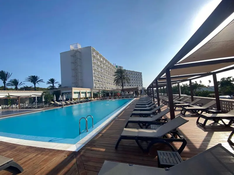 Une piscine turquoise bordée de transats sous un auvent en bois, avec un bâtiment hôtelier en arrière-plan.