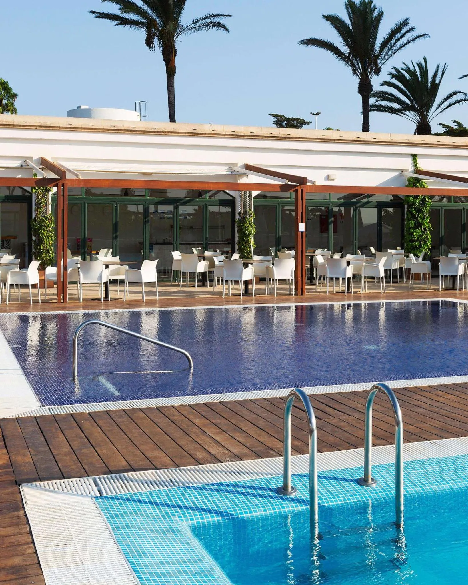 Une piscine rectangulaire avec des escaliers d'accès et une terrasse ombragée avec des tables et des chaises.