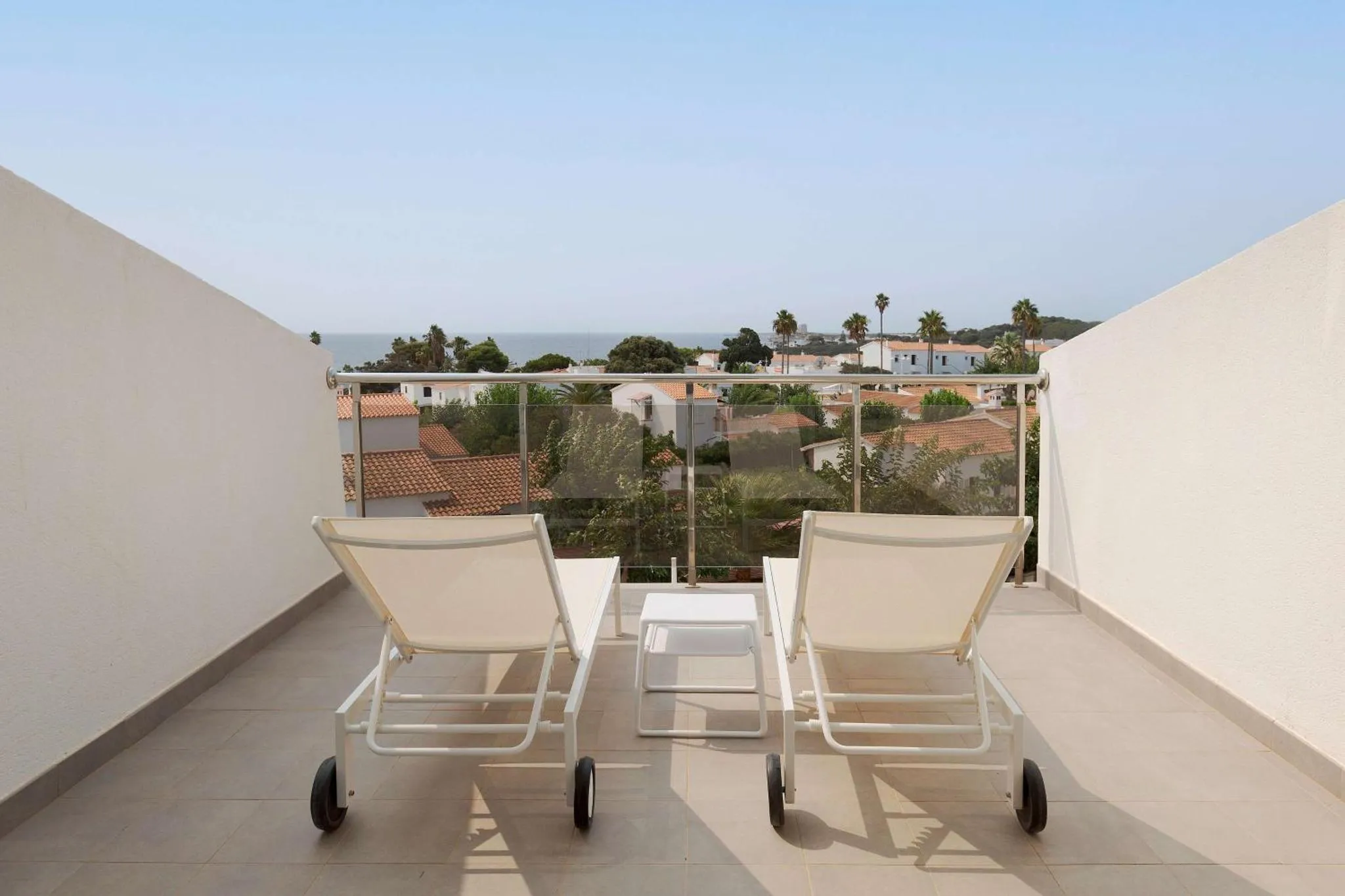Deux transats blancs sur une terrasse ensoleillée avec vue sur la mer et la végétation.