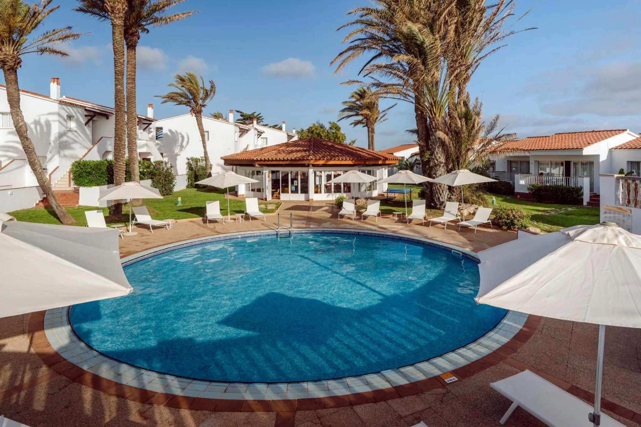 Piscine rectangulaire entourée de chaises longues et de parasols blancs, dans un jardin tropical.