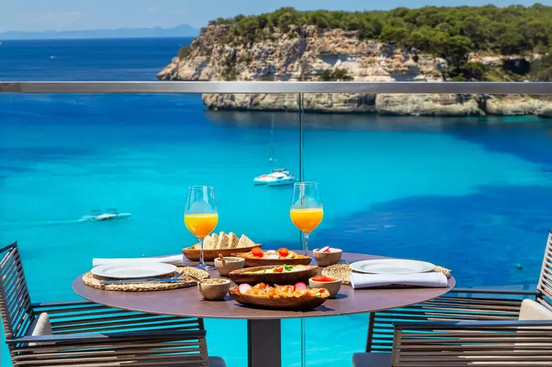 Une table est dressée avec des couverts et des boissons sur une terrasse offrant une vue sur la mer.