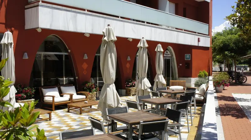 Terrasse ombragée avec tables et canapés devant un bâtiment rouge et blanc.