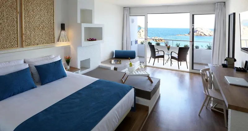 Chambre lumineuse avec un grand lit et une vue sur la mer depuis un balcon.