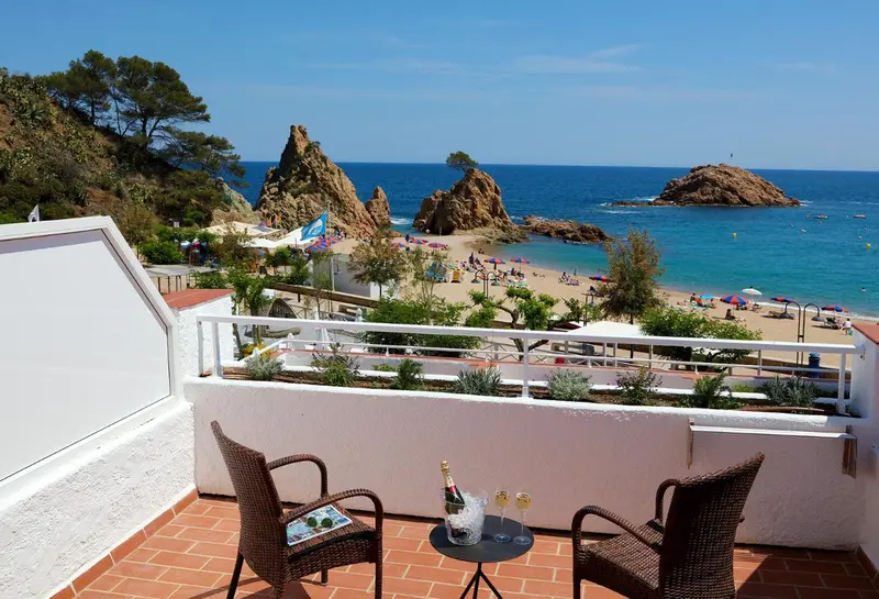 Deux fauteuils et une table basse sur une terrasse ensoleillée avec vue sur une plage et des formations rocheuses.