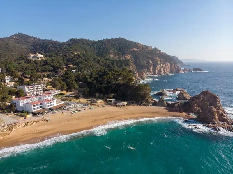 Vue aérienne d'une plage de sable fin bordée de bâtiments blancs et de collines verdoyantes.