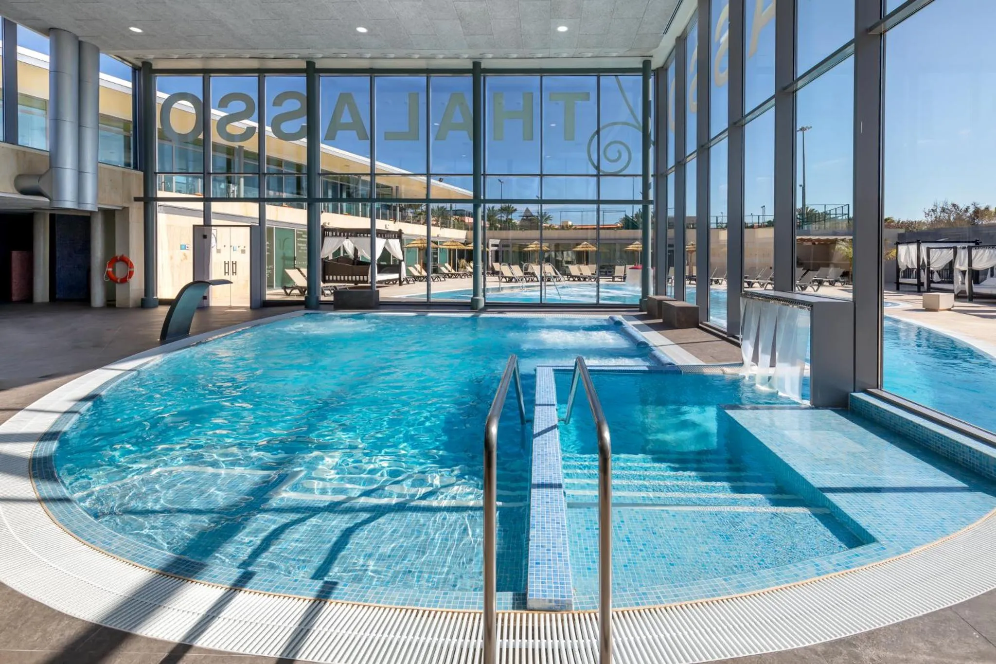Une piscine intérieure avec des transats et des palmiers visibles à travers de grandes baies vitrées.