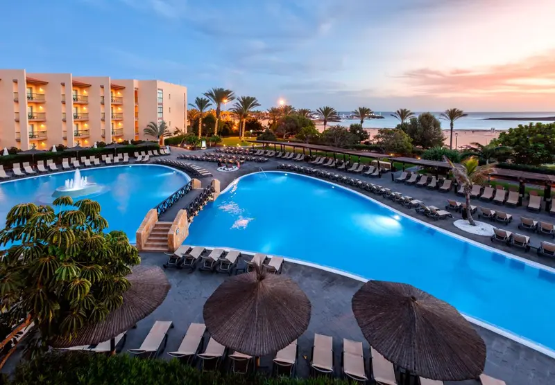 Une grande piscine turquoise bordée de transats et de parasols, avec un bâtiment hôtelier en arrière-plan.