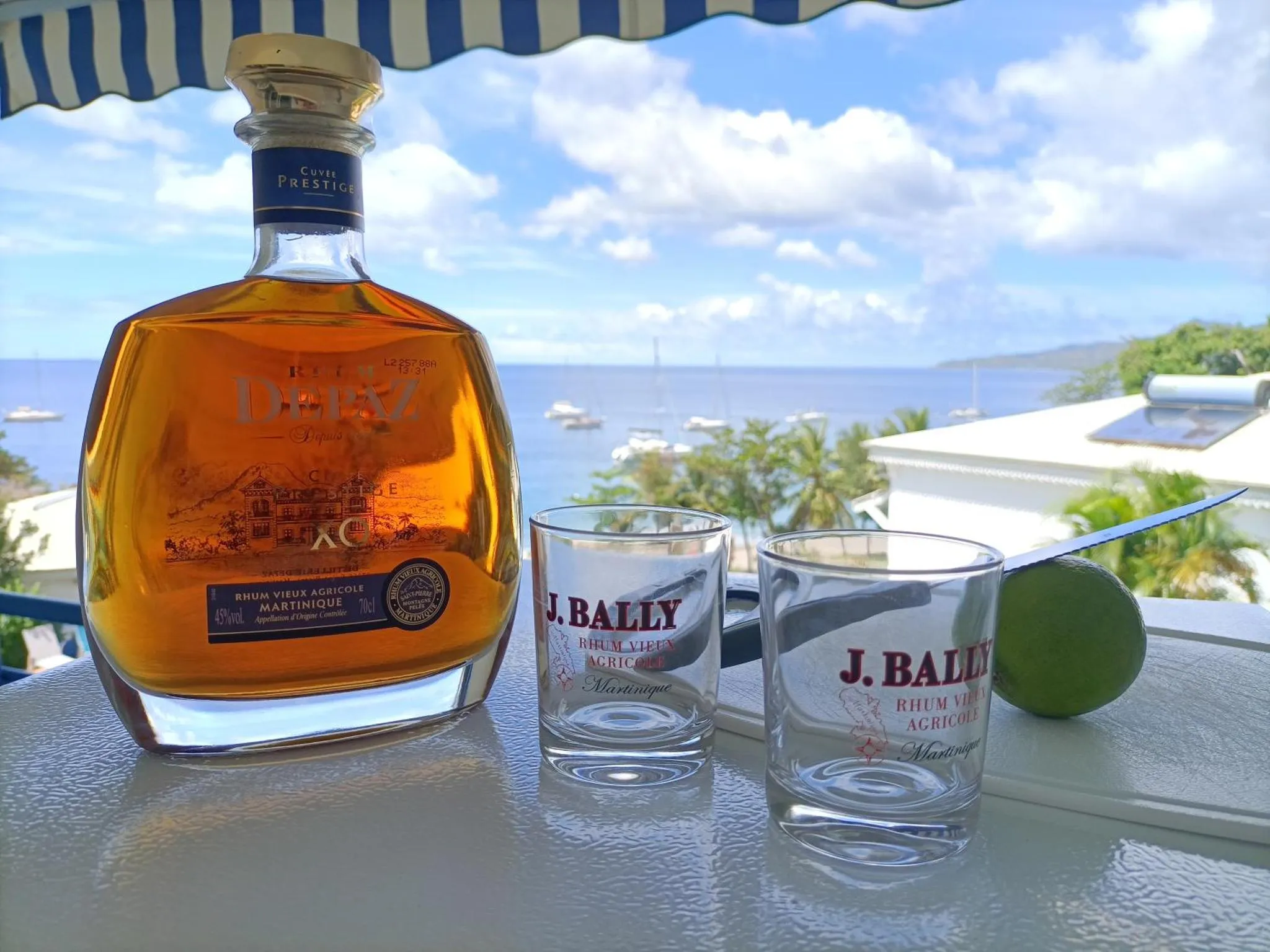 Une bouteille de rhum et deux verres sur une table blanche avec vue sur la mer et les collines.