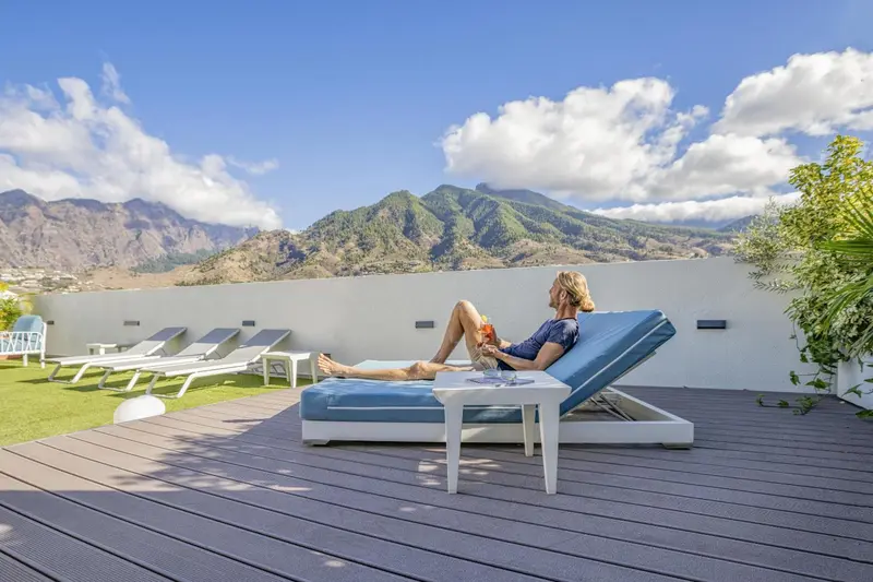 Une personne se relaxe sur un transat au bord d'une terrasse ensoleillée avec vue sur des montagnes.
