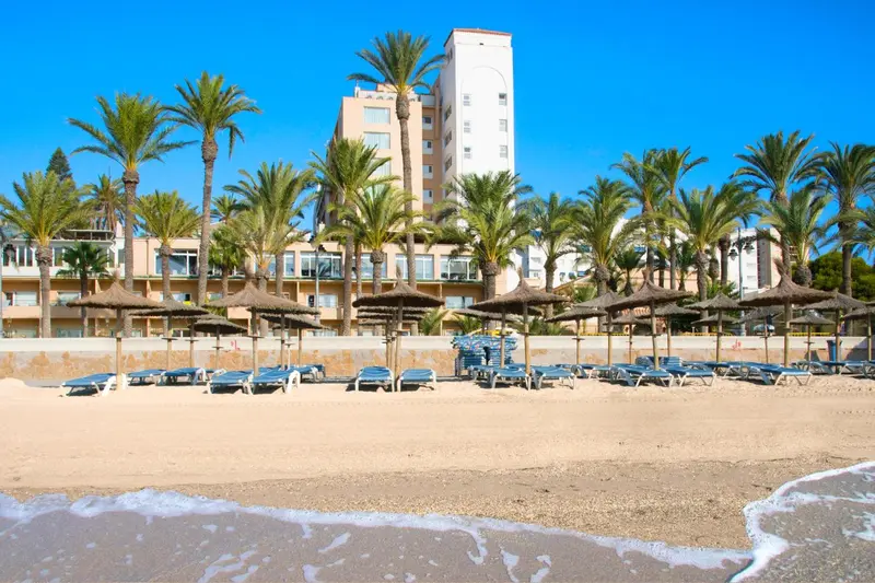 Plage de sable fin bordée de palmiers et de transats bleus devant un bâtiment blanc.