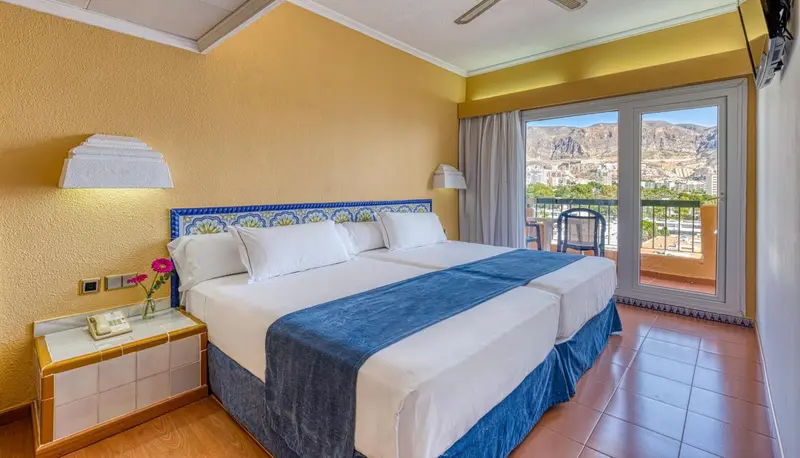 Une chambre lumineuse avec un grand lit et un balcon offrant une vue sur les montagnes.