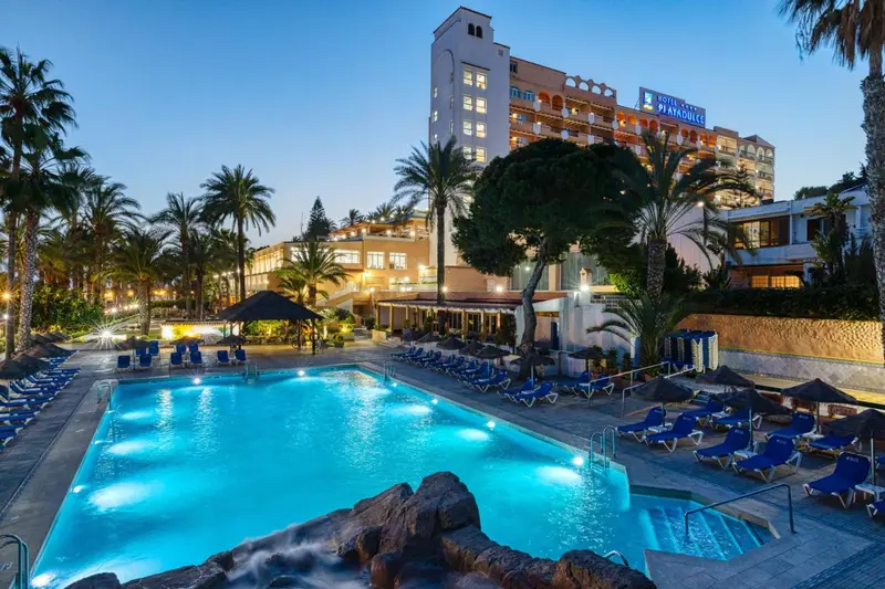 Vue d'un hôtel illuminé avec une piscine au premier plan et des palmiers.