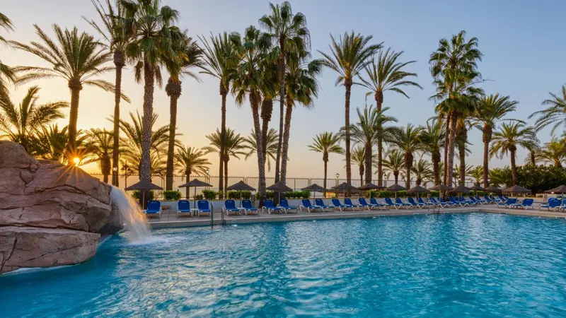 Une piscine turquoise bordée de transats, avec des palmiers et une cascade rocheuse au premier plan.