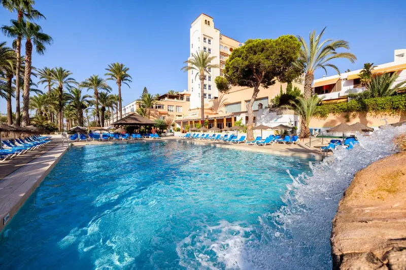 Une piscine turquoise bordée de transats, avec un bâtiment beige et des palmiers en arrière-plan.