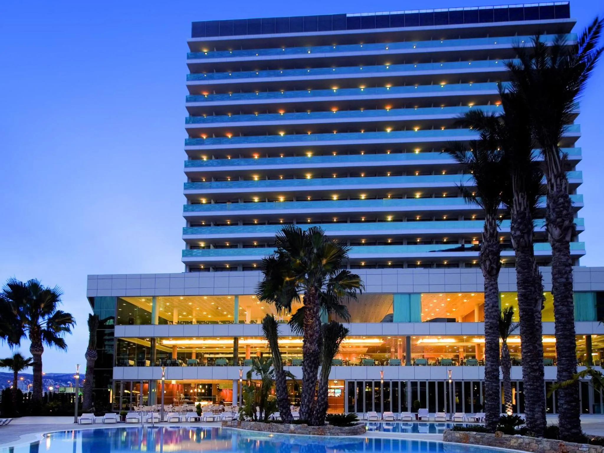 Bâtiment moderne illuminé au crépuscule, bordé de palmiers et surplombant une piscine.