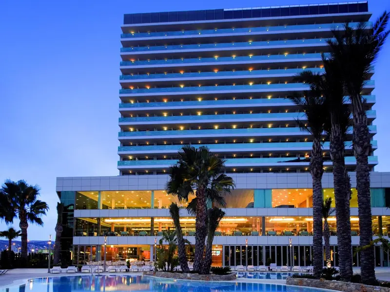 Bâtiment moderne illuminé au crépuscule, bordé de palmiers et surplombant une piscine.