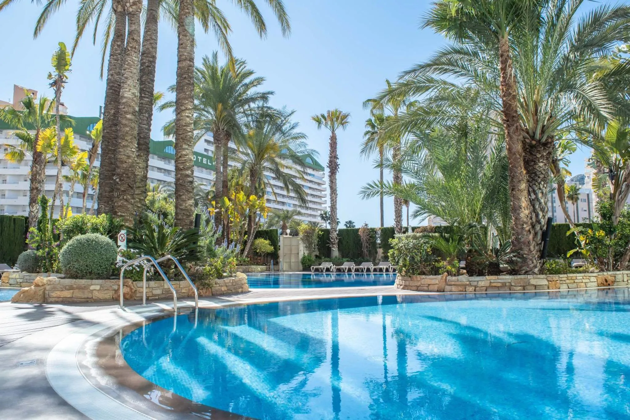 Une piscine turquoise bordée de palmiers et de transats, devant un bâtiment hôtelier.