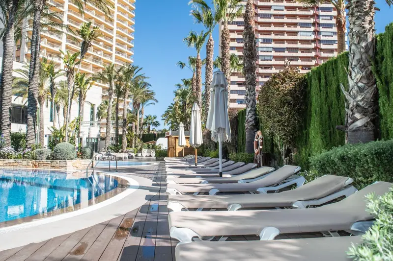 Transats alignés au bord d'une piscine ensoleillée, avec des bâtiments en arrière-plan.