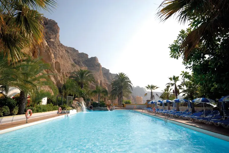 Une piscine rectangulaire bordée de transats, avec une cascade artificielle et des palmiers.