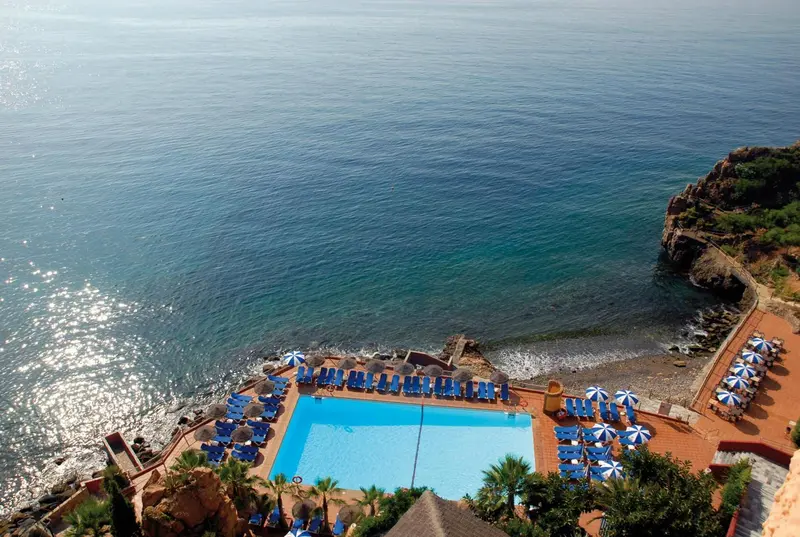 Vue aérienne d'une piscine et d'une plage rocheuse bordée de transats bleus.
